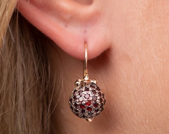 May include: A pair of gold earrings with a round, red gemstone design. The earrings are dangling from the earlobe.