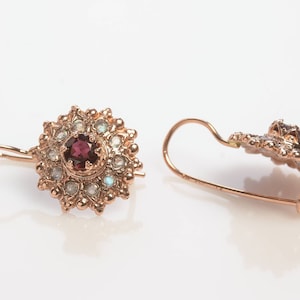 May include: A pair of gold earrings with a red gemstone surrounded by smaller white gemstones. The earrings have a delicate, floral design.