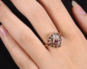 May include: A gold ring with a heart-shaped design set with red gemstones. The ring is worn on the finger of a person's hand.