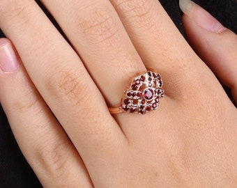May include: A rose gold ring featuring a cluster of small, round, red gemstones. The ring's design is ornate, with the gemstones arranged in a floral or geometric pattern. The ring is on a person's finger, showcasing the jewelry.