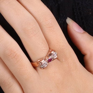 Op de afbeelding: Een ring van roségoud met een strikdesign met een rode edelsteen in het midden en heldere edelstenen rond de strik.