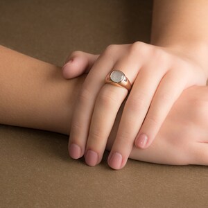 May include: A gold ring with a white oval gemstone. The ring is on a person's finger.