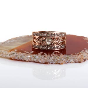 May include: A rose gold ring with a large, round, clear gemstone surrounded by smaller, clear gemstones. The ring is set on a brown agate slice.