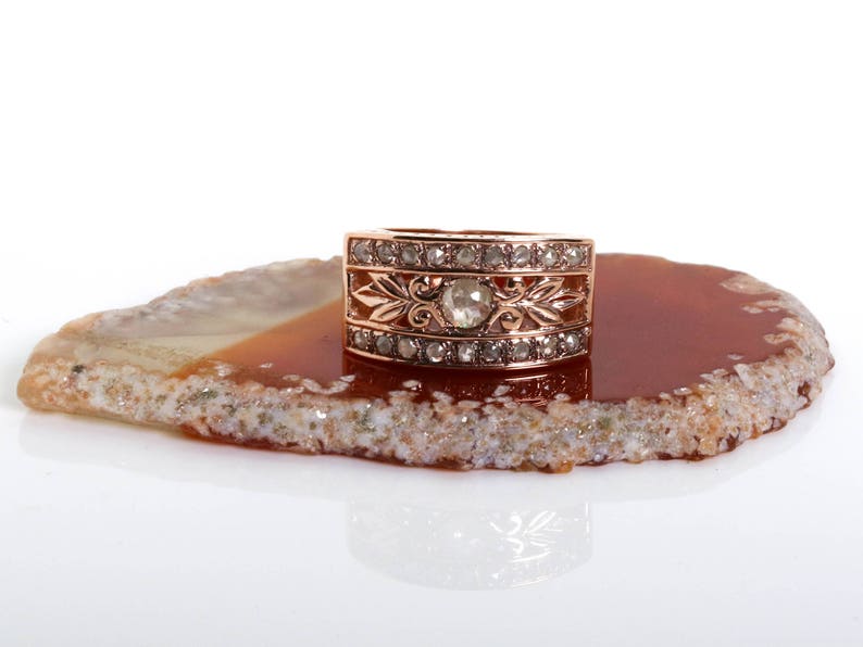 May include: A rose gold ring with a large, round, clear gemstone surrounded by smaller, clear gemstones. The ring is set on a brown agate slice.