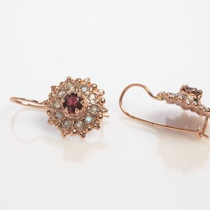 May include: A pair of gold earrings with a red gemstone surrounded by smaller white gemstones. The earrings have a delicate floral design.