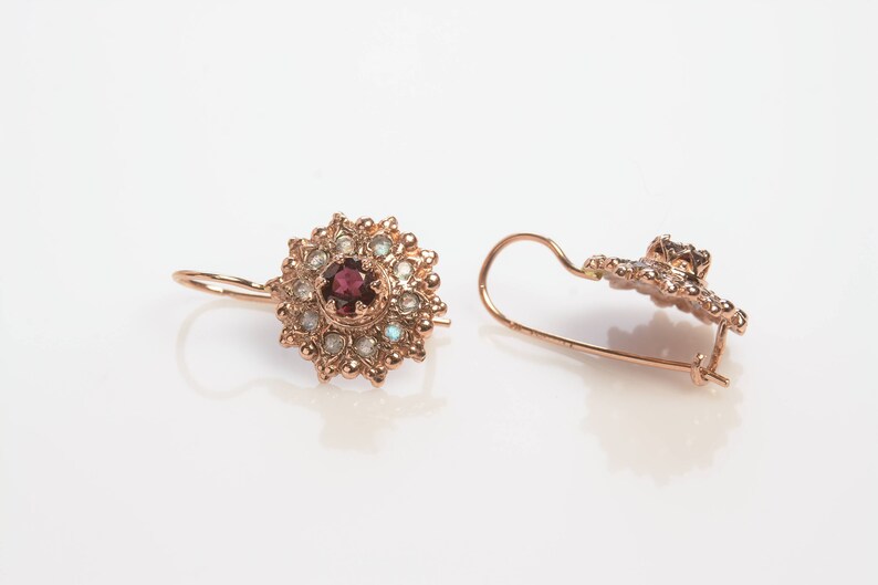 May include: A pair of gold earrings with a red gemstone surrounded by smaller white gemstones. The earrings have a delicate floral design.
