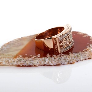 May include: A rose gold ring with a band of small diamonds and a larger diamond in the center. The ring is sitting on a brown and white agate slice.