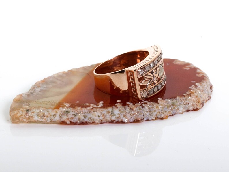 May include: A rose gold ring with a band of small diamonds and a larger diamond in the center. The ring is sitting on a brown and white agate slice.