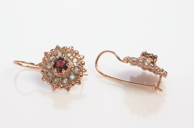 May include: A pair of gold earrings with a red gemstone surrounded by smaller white gemstones. The earrings have a delicate, floral design.