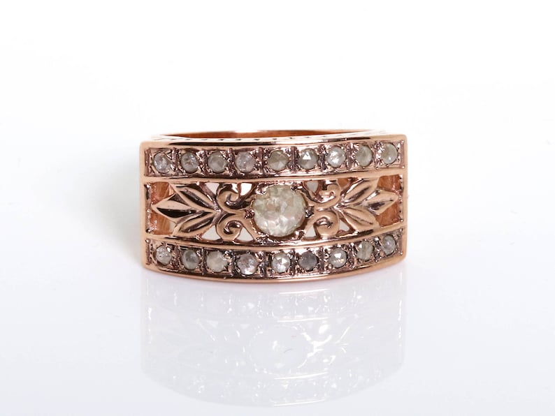 May include: A rose gold ring with a large, clear, round gemstone surrounded by smaller, clear gemstones. The ring has a decorative, ornate design.