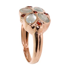 May include: A rose gold ring with five oval-shaped gemstones. The center stone is larger and surrounded by four smaller stones. The smaller stones are red, and the larger stone is a light blue color.