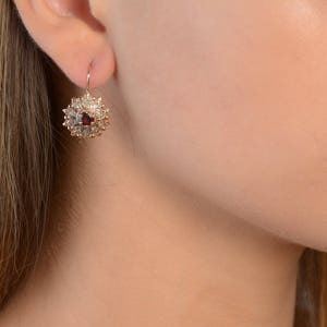 May include: A pair of gold earrings with a red gemstone surrounded by smaller clear gemstones in a floral design.