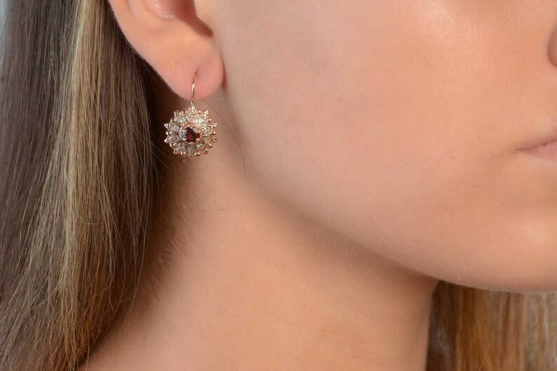 May include: A pair of gold earrings with a red gemstone surrounded by smaller clear gemstones in a floral design.