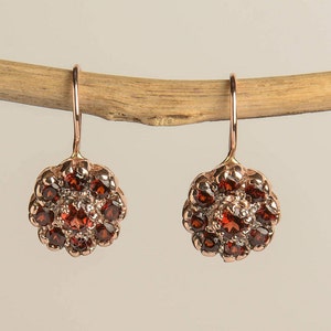 May include: A pair of dangle earrings with a rose gold finish. Each earring features a cluster of small, round, red gemstones set in a flower-shaped design.