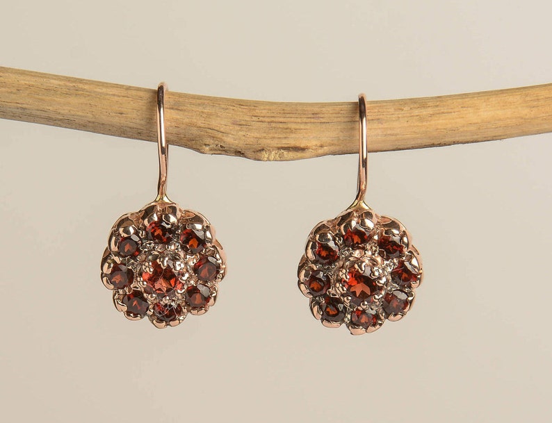 May include: A pair of dangle earrings with a rose gold finish. Each earring features a cluster of small, round, red gemstones set in a flower-shaped design.