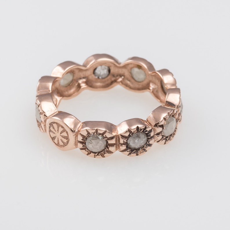 May include: A rose gold band ring with a row of round, gray diamonds set in a sunburst design.