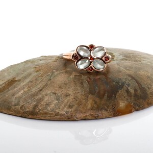 May include: A rose gold ring with a flower-shaped design featuring five clear stones and red accents.