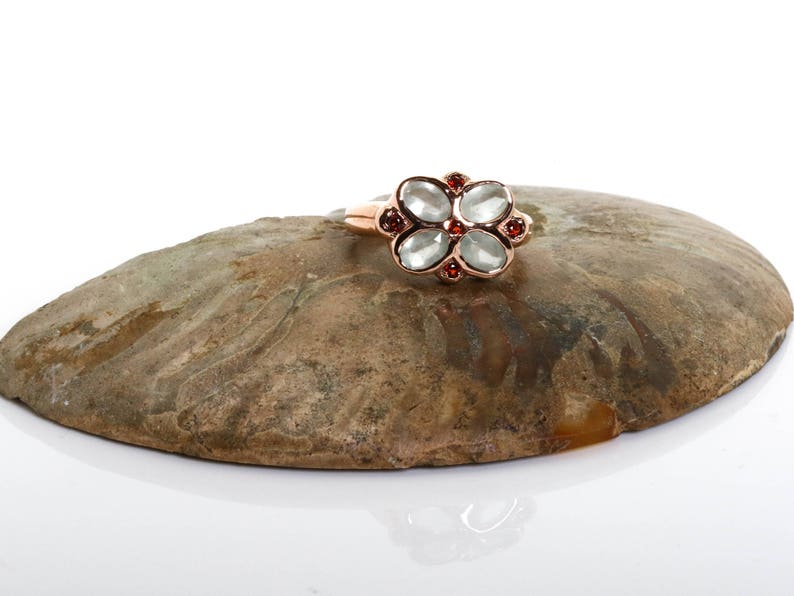 May include: A rose gold ring with a flower-shaped design featuring five clear stones and red accents.