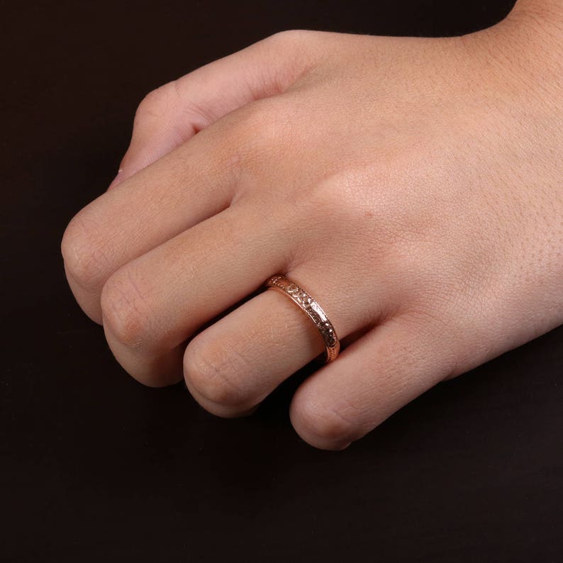 May include: A rose gold band ring with a textured design. The ring is worn on the ring finger of a hand.