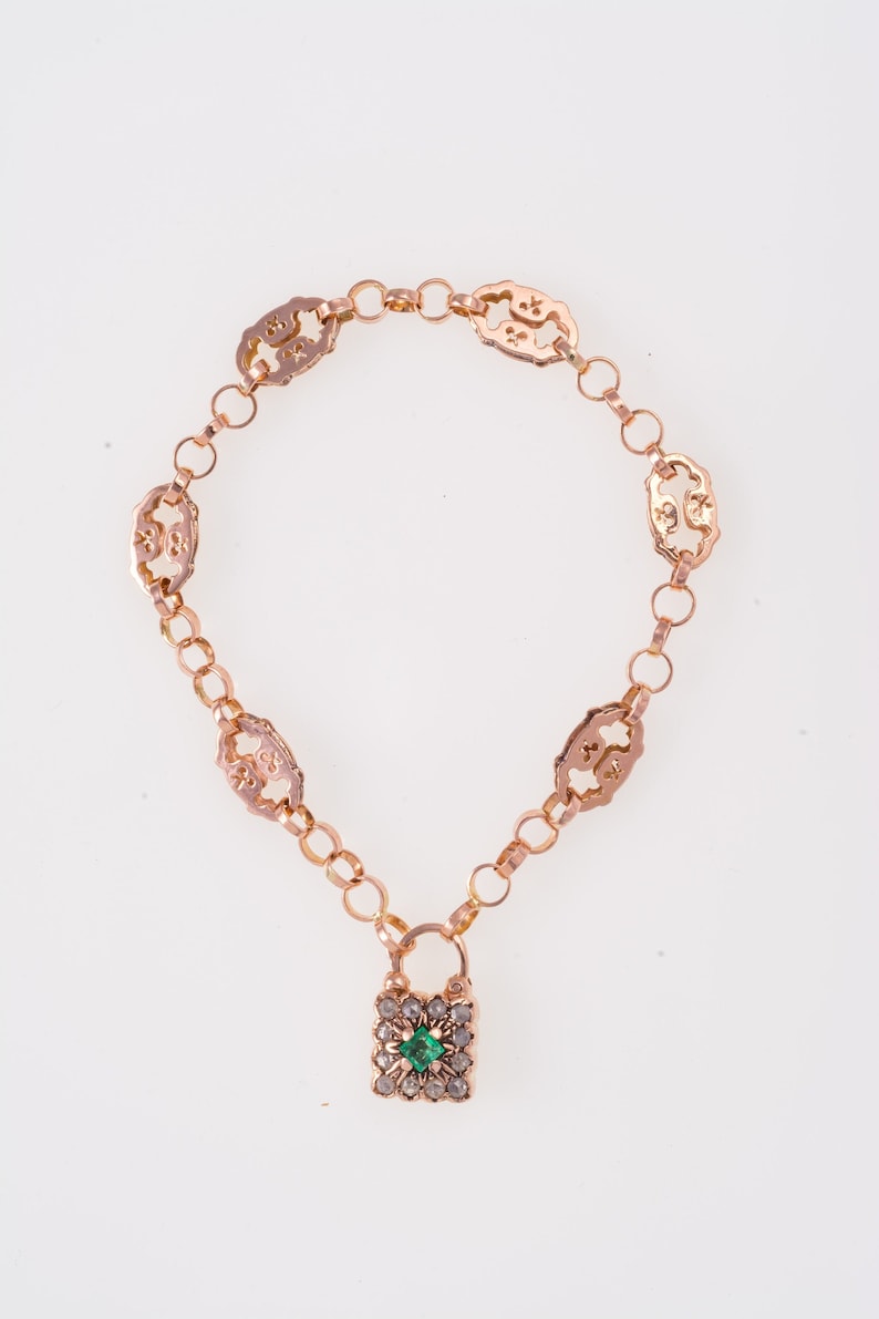 Op de afbeelding: Een gouden kettingarmband met een vierkante hanger met een groene edelsteen omringd door kleinere heldere edelstenen.