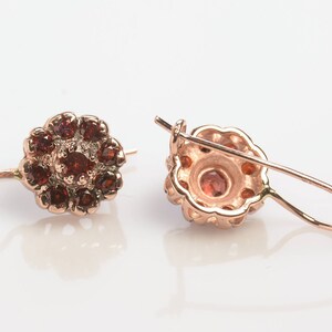 May include: A pair of rose gold earrings with a flower design. The earrings are adorned with small, red gemstones.