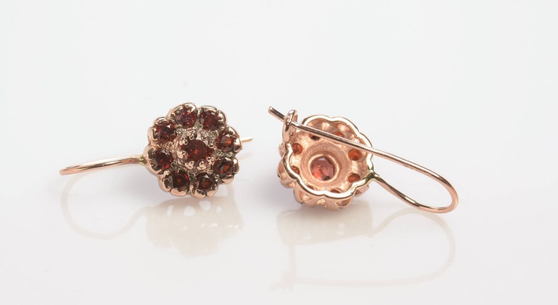 May include: A pair of rose gold earrings with a flower design. The earrings are adorned with small, red gemstones.