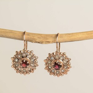 May include: Rose gold dangle earrings with a red gemstone surrounded by smaller white gemstones in a circular design.