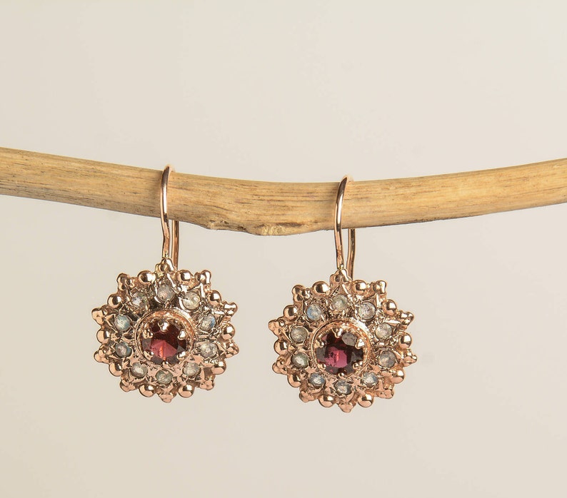 May include: Rose gold dangle earrings with a red gemstone surrounded by smaller white gemstones in a circular design.