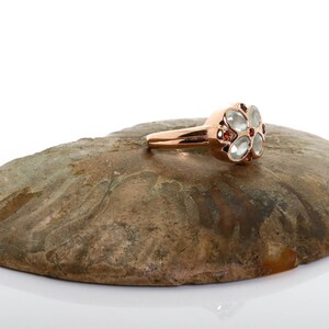 May include: A rose gold ring with a flower-shaped design. The center of the flower is made of clear stones, and the petals are accented with small red stones.