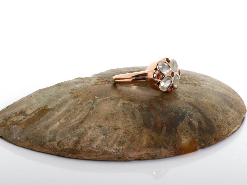 May include: A rose gold ring with a flower-shaped design. The center of the flower is made of clear stones, and the petals are accented with small red stones.
