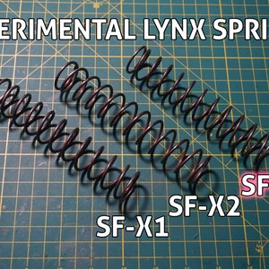 Puede incluir: Tres muelles metálicos etiquetados como "SF-X1", "SF-X2" y "SF-X3" sobre una estera de corte verde. Los muelles están enrollados y parecen ser para un dispositivo mecánico.