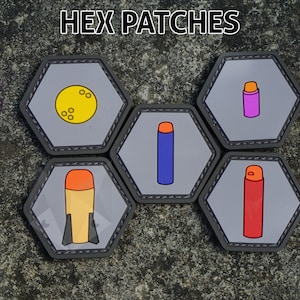 Pode incluir: Cinco patches hexagonais com diferentes designs. Os patches são cinzentos com costuras pretas ao redor das bordas. Os designs incluem um círculo amarelo com pontos pretos, um tubo azul com uma parte superior laranja, um tubo vermelho com uma parte superior laranja, um tubo rosa com uma parte superior laranja e um foguete amarelo e laranja com barbatanas pretas.
