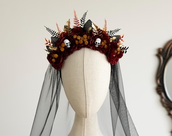Puede incluir: Un tocado con flores artificiales de color rojo oscuro, pequeñas calaveras blancas y follaje negro y dorado similar a un helecho. Un velo largo de tul negro transparente está sujeto a la base del arreglo floral y cae hacia los lados.