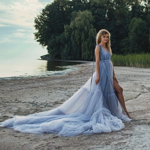 May include: A woman wearing a light blue tulle gown with a long train, standing on a sandy beach with a body of water in the background.