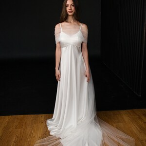 Puede incluir: Un vestido de novia de sat&eacute;n blanco con una superposici&oacute;n de tul y una larga cola. El vestido tiene un dise&ntilde;o simple y elegante con un escote de pico y tirantes finos. La superposici&oacute;n de tul agrega un toque de romance y fantas&iacute;a al vestido.