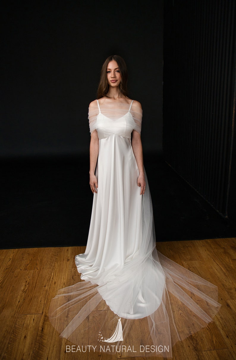 Puede incluir: Un vestido de novia de sat&eacute;n blanco con una superposici&oacute;n de tul y una larga cola. El vestido tiene un dise&ntilde;o simple y elegante con un escote de pico y tirantes finos. La superposici&oacute;n de tul agrega un toque de romance y fantas&iacute;a al vestido.