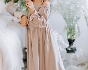 May include: A young girl in a light brown dress holds a white flower. She is standing in a room with white flowers in the background.