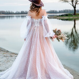 A bride wearing a white tulle wedding dress with a brown hat and a bouquet of dried flowers. The dress has a lace-up back and a fitted bodice. The bride is standing on a sandy beach with a lake in the background.