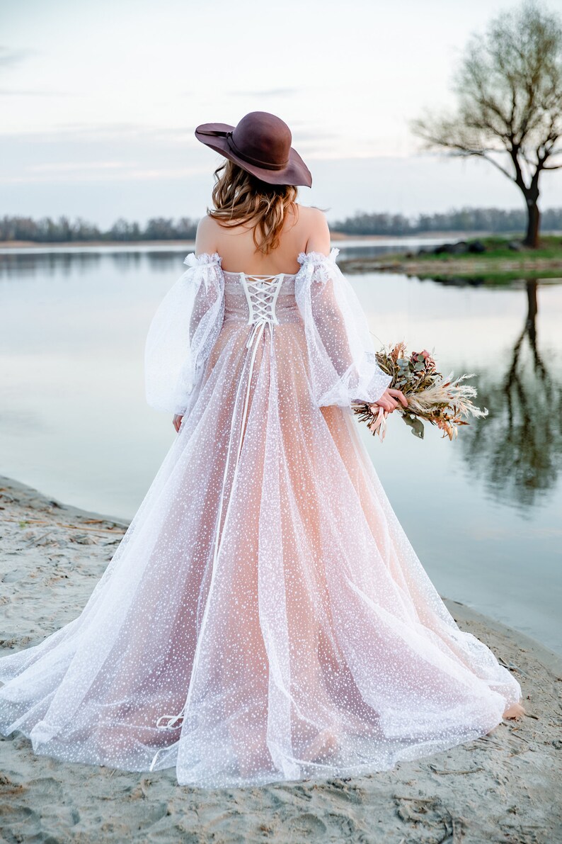 A bride wearing a white tulle wedding dress with a brown hat and a bouquet of dried flowers. The dress has a lace-up back and a fitted bodice. The bride is standing on a sandy beach with a lake in the background.