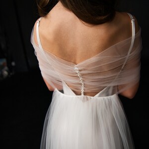Puede incluir: Un vestido de novia de tul blanco con la espalda descubierta y una fila de botones de perla. El vestido tiene una falda fluida y un corpi&ntilde;o ajustado. El vestido est&aacute; dise&ntilde;ado por Beauty Natural Design.