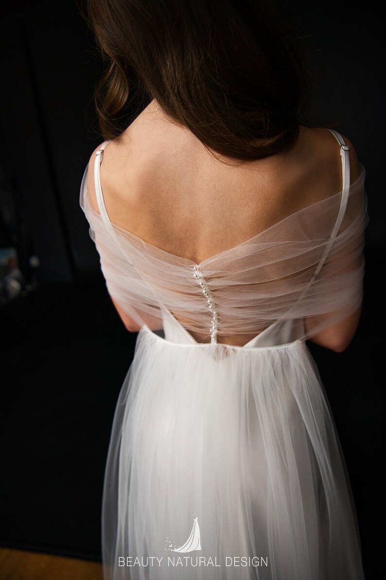 Puede incluir: Un vestido de novia de tul blanco con la espalda descubierta y una fila de botones de perla. El vestido tiene una falda fluida y un corpi&ntilde;o ajustado. El vestido est&aacute; dise&ntilde;ado por Beauty Natural Design.