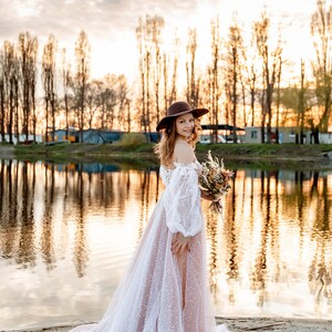 A woman in a white lace wedding dress with a brown hat and a bouquet of flowers stands on a sandy beach by a lake at sunset.
