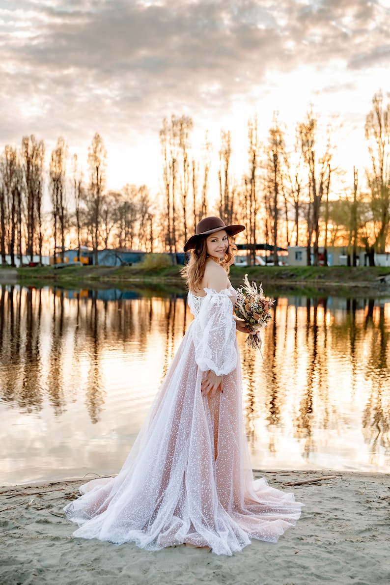 A woman in a white lace wedding dress with a brown hat and a bouquet of flowers stands on a sandy beach by a lake at sunset.