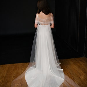 Puede incluir: Un vestido de novia de tul blanco con una superposici&oacute;n transparente y una cola larga. El vestido tiene la espalda baja y un dise&ntilde;o sencillo.