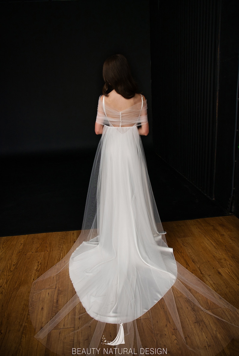 Puede incluir: Un vestido de novia de tul blanco con una superposici&oacute;n transparente y una cola larga. El vestido tiene la espalda baja y un dise&ntilde;o sencillo.