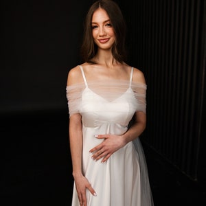 Puede incluir: Una mujer con un vestido de novia blanco de hombros descubiertos con una superposici&oacute;n de tul. Est&aacute; de pie frente a un fondo negro y sonr&iacute;e a la c&aacute;mara. El vestido tiene un dise&ntilde;o simple y elegante, perfecto para una novia moderna.