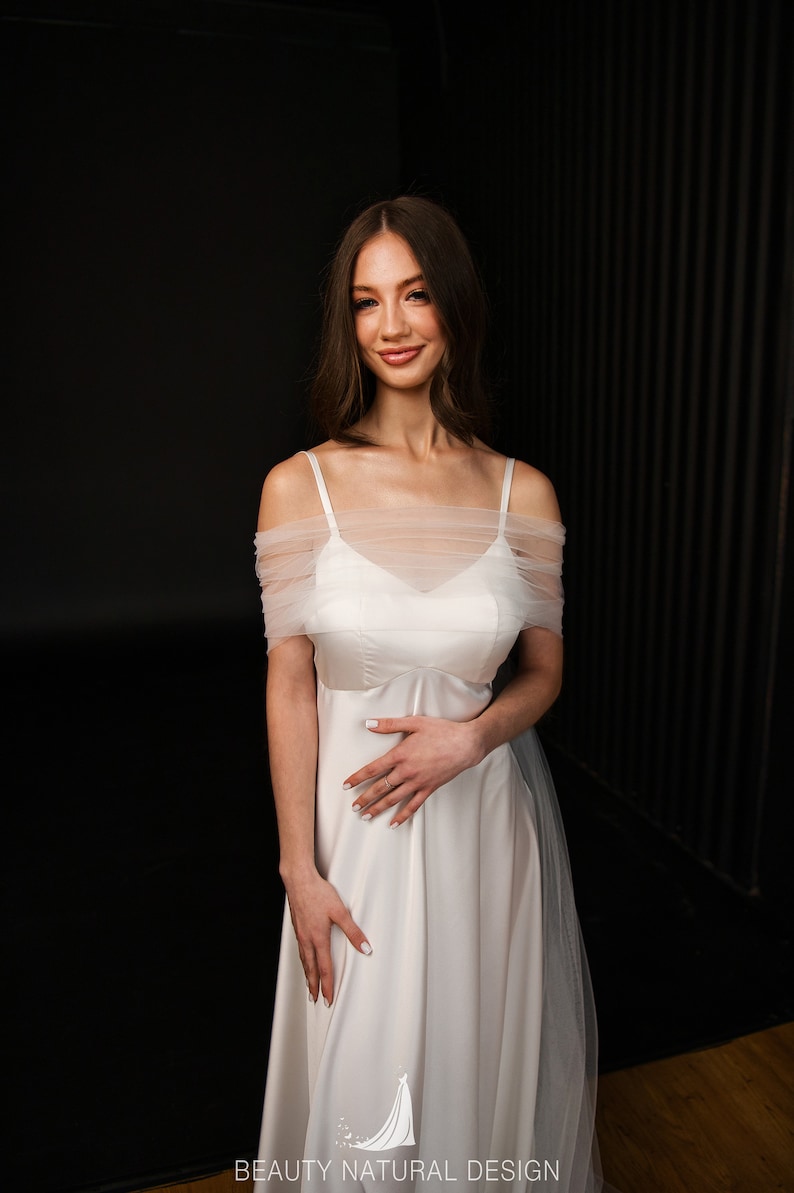 Puede incluir: Una mujer con un vestido de novia blanco de hombros descubiertos con una superposici&oacute;n de tul. Est&aacute; de pie frente a un fondo negro y sonr&iacute;e a la c&aacute;mara. El vestido tiene un dise&ntilde;o simple y elegante, perfecto para una novia moderna.