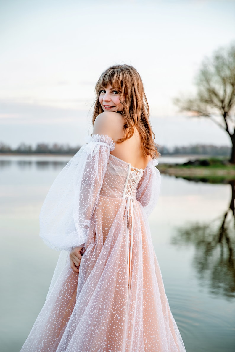 A woman wearing a white off-the-shoulder dress with a lace bodice and a tulle skirt. The dress is covered in small white dots. She is standing in front of a body of water with a tree in the background.