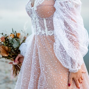 This beige off-the-shoulder wedding dress features a lace bodice and a tulle skirt with white polka dots. The dress has long, puffy sleeves.