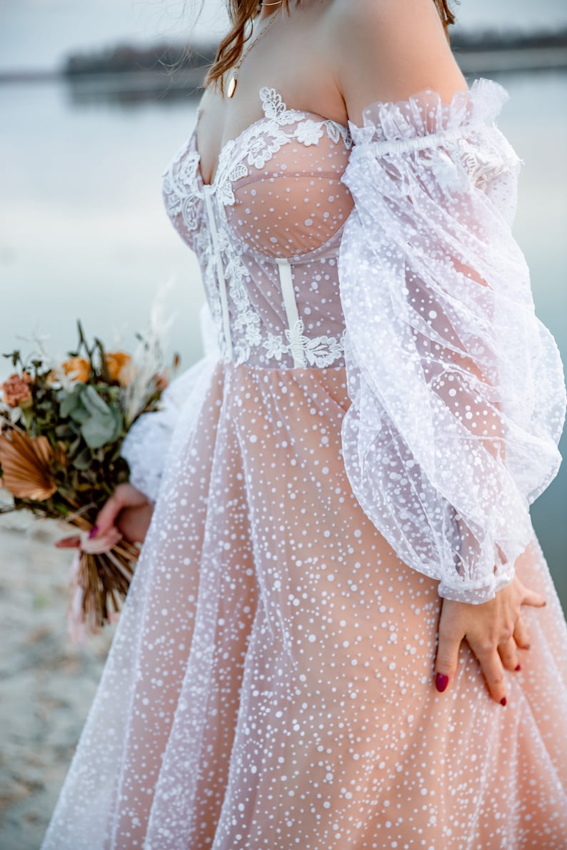 This beige off-the-shoulder wedding dress features a lace bodice and a tulle skirt with white polka dots. The dress has long, puffy sleeves.